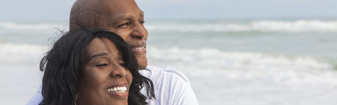 Middle aged black couple holding each other and smiling with white teeth on the beach