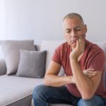 Photo of Depressed ill man having toothache and touching cheek. Mature man suffering from tooth pain, caries. Handsome gray hair male suffering from toothache, closeup. Portrait of casual 46 years mature man toothache with painful expression, sitting on sofa at home,