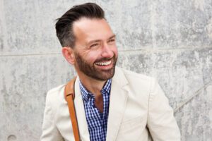 cheerful middle aged man with beard standing against wall and smiling